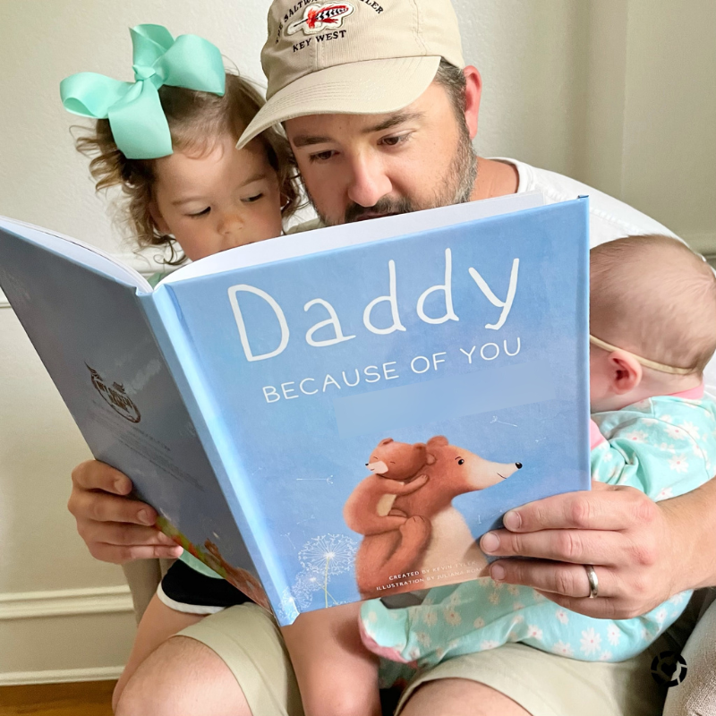 Dad sitting with a toddler girl and baby reading a book called Daddy Because of You for Father's Day